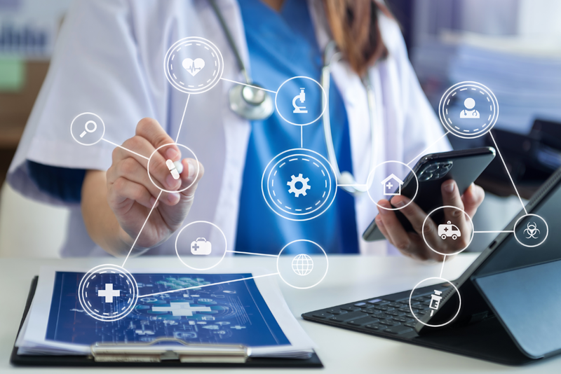 A healthcare professional in scrubs pointing a digital stylus toward a floating network of medical icons, with a smartphone in hand and a tablet on the desk.