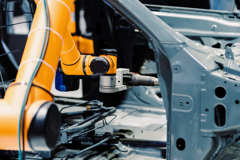 An automated bright orange robotic arm precision-welding or assembling the interior frame of a grey car chassis on an automotive factory production line.