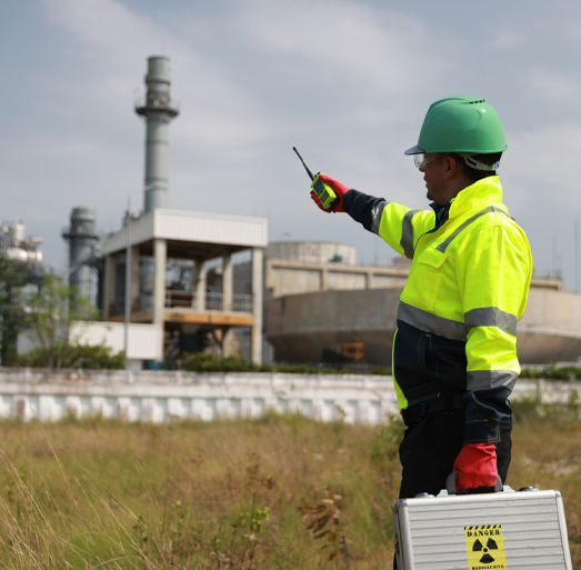 A site operator wearing a bright yellow high-visibility jacket, green hard hat, and red gloves stands in a grassy field holding a walkie-talkie and pointing toward an industrial facility in the background. In the foreground, he holds a silver briefcase marked with a yellow radioactive danger symbol.
