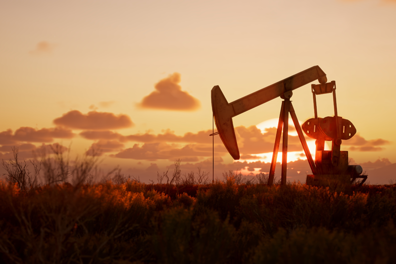 Silhouette of an oil pumpjack operating in a field during a vibrant orange sunset with scattered clouds in the background.