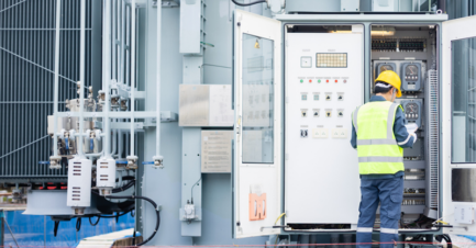 An electrical engineer wearing a yellow hard hat and high-visibility safety vest standing in front of an open outdoor electrical control panel, reviewing a clipboard at a power facility.