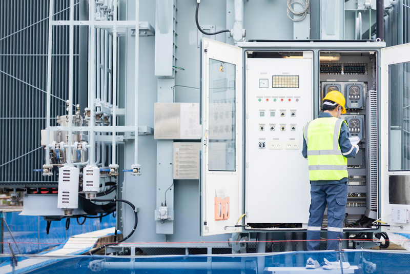 An electrical engineer wearing a yellow hard hat and high-visibility safety vest standing in front of an open outdoor electrical control panel, reviewing a clipboard at a power facility.