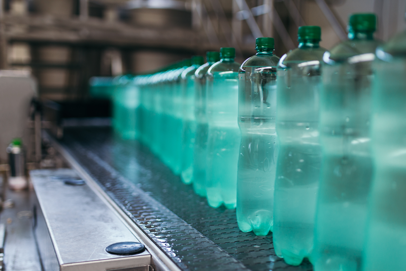 A long row of green plastic beverage bottles filled with clear liquid moving along an automated conveyor belt system in a manufacturing plant.