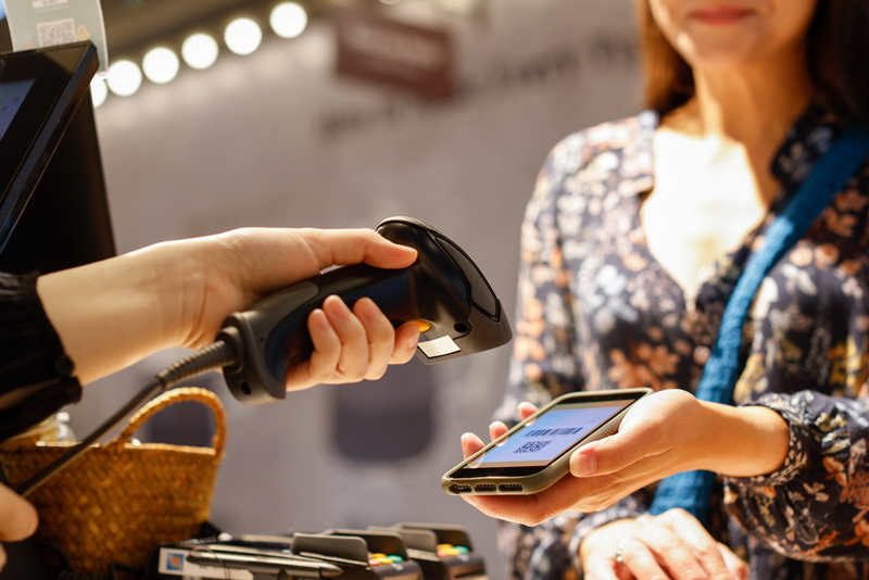 Close-up of a store cashier using a handheld barcode scanner to scan a QR code digital payment from a customer’s smartphone screen at a retail checkout counter.